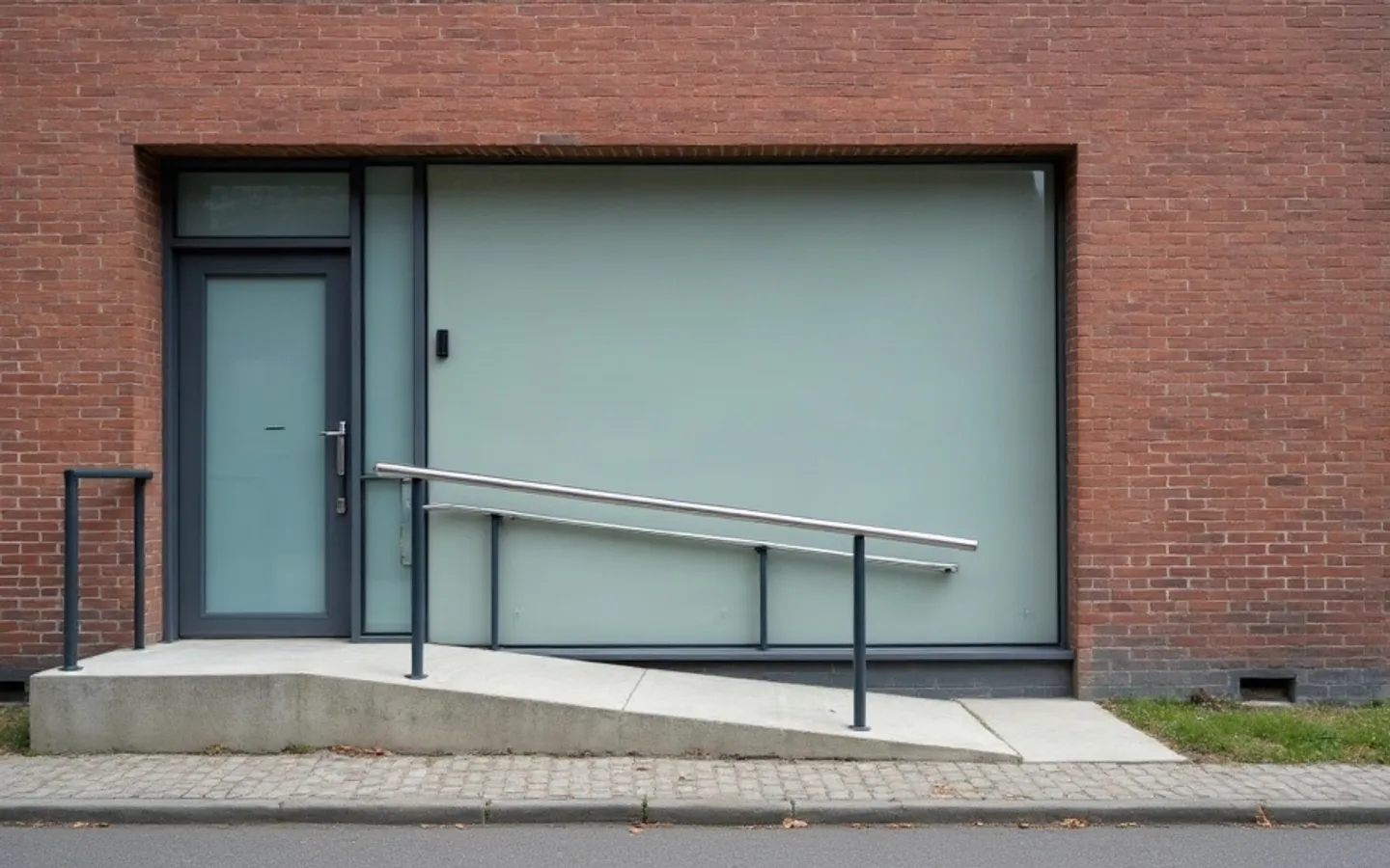Zij-ingang van een bakstenen woonhuis met een deur van matglas en een betonnen hellingbaan met stalen leuning. Naast de deur is op de muur een lichte rechthoek zichtbaar waar een naambord heeft gehangen.