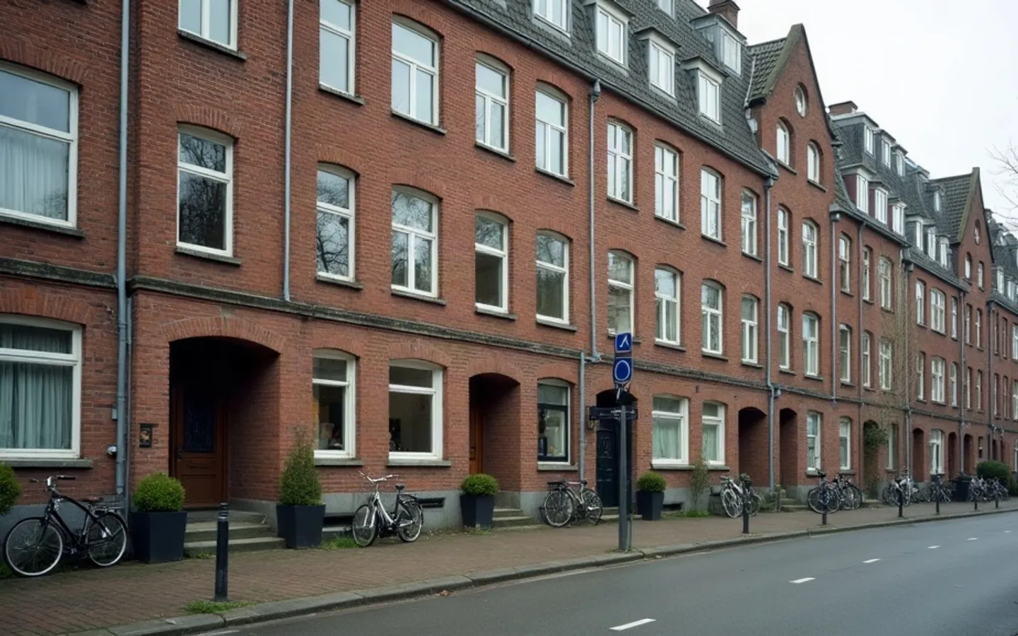 Rij bakstenen rijtjeshuizen in een Nederlandse straat in zacht ochtendlicht. Bij één woning zijn de gordijnen weg en de ramen kaal, bij de buurhuizen staan planten en lampen achter het glas.